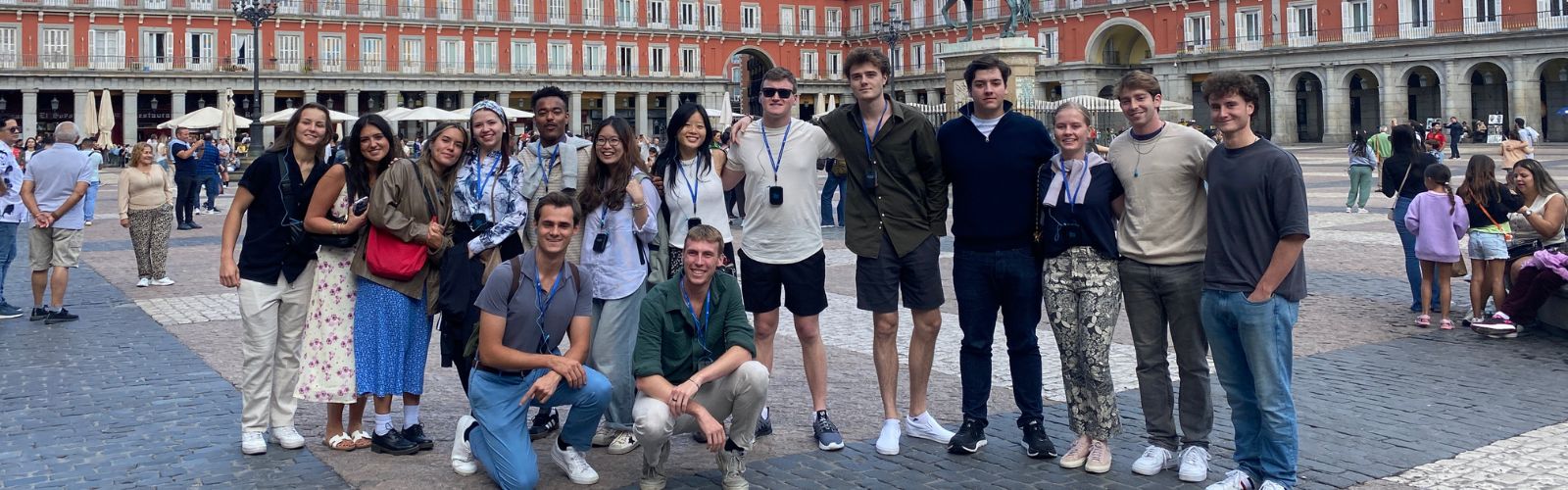 Fifteen students posed in a large plaza surrounded by a red building.